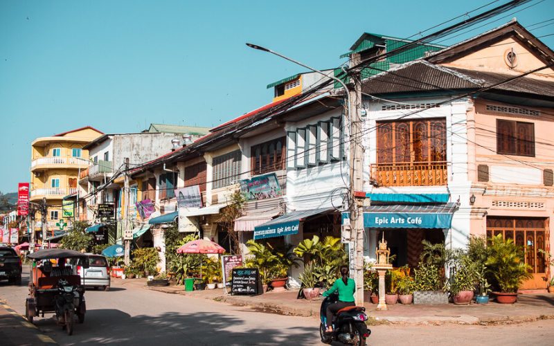 Rue animée de Kampot, bordée de maisons coloniales françaises, de cafés locaux et de petites boutiques, avec un tuk-tuk et des motos circulant paisiblement sous un ciel ensoleillé