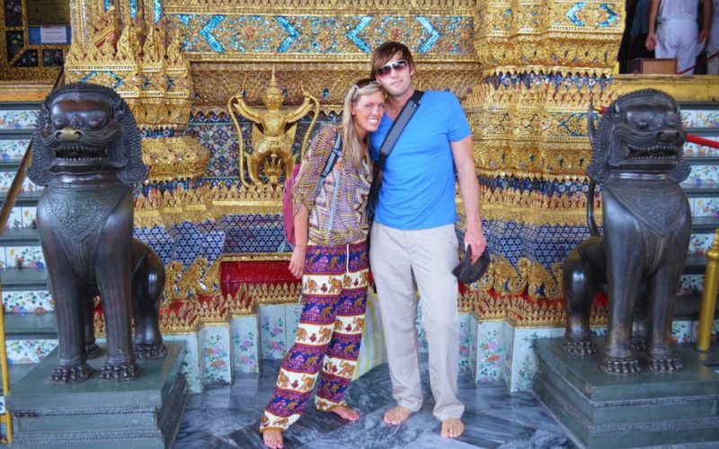Deux touristes posent pieds nus devant un temple thaï orné de sculptures dorées et gardé par des statues de lions mythiques.