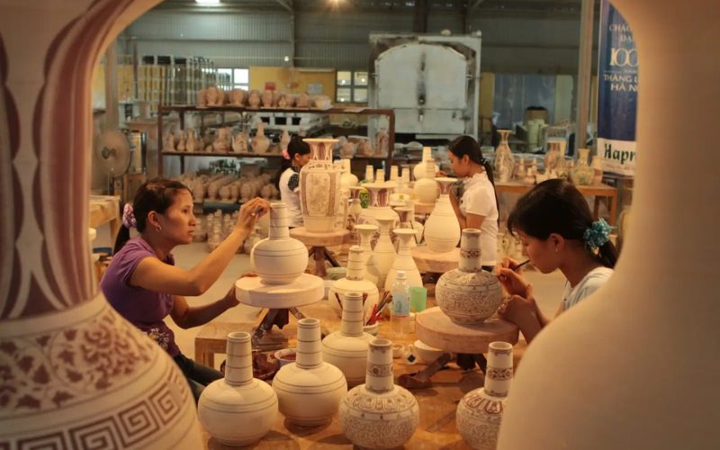 Alt text (French) :Artisanes du village de céramique de Chu Dau, à Hai Duong, décorant minutieusement des vases en argile. Les poteries blanches aux motifs délicats témoignent du savoir-faire ancestral et raffiné de ce village artisanal vietnamien renommé