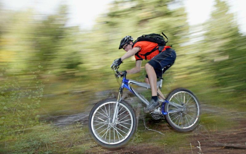 Homme en VTT dévalant un sentier forestier à toute vitesse, équipé d’un casque, de gants et d’un sac à dos, illustrant une descente intense en pleine nature