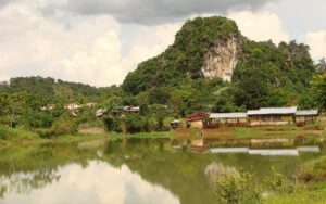 Village rural du Nord Laos avec des maisons traditionnelles au pied d’une colline calcaire recouverte de végétation, se reflétant dans un paisible étang sous un ciel nuageux