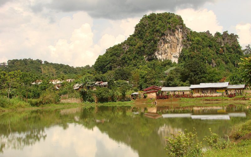 Village rural du Nord Laos avec des maisons traditionnelles au pied d’une colline calcaire recouverte de végétation, se reflétant dans un paisible étang sous un ciel nuageux