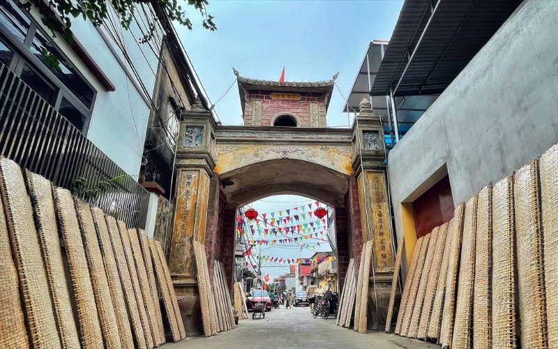 Porte ancienne du village de Thổ Hà, à Bắc Giang, avec des claies de bambou couvertes de galettes de riz mises à sécher le long des murs, et des fanions colorés décorant la rue animée
