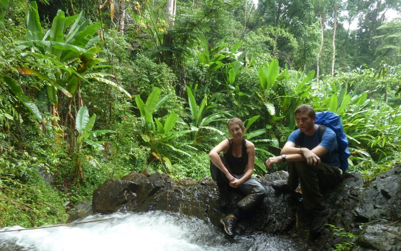 Deux randonneurs assis sur un rocher au bord d’un ruisseau dans une jungle tropicale dense, entourés de végétation luxuriante et de grandes feuilles de bananiers