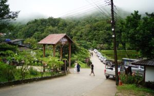Entrée du village de Pilok, entourée de montagnes verdoyantes et de brume, avec route bordée de voitures et quelques visiteurs à pied