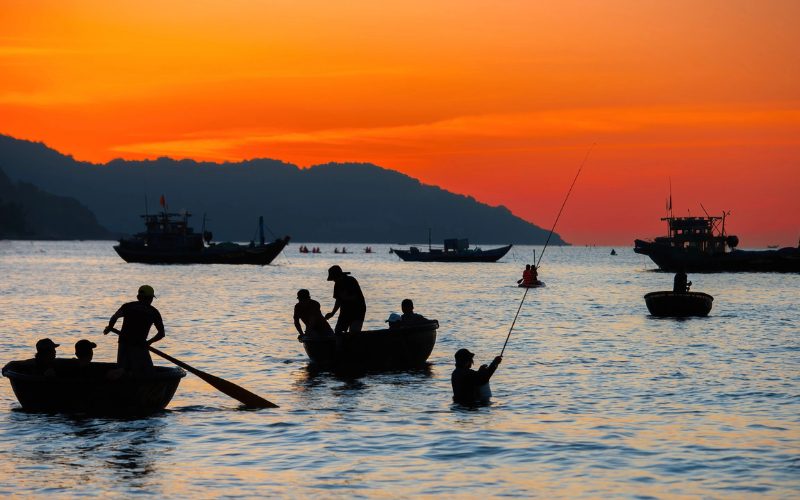 Silhouettes de pêcheurs vietnamiens dans des bateaux paniers ronds au coucher du soleil, sur une mer calme aux reflets argentés, avec plusieurs bateaux de pêche au large et des collines sombres se détachant sous un ciel orange flamboyant