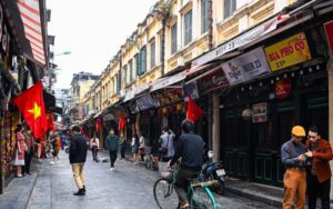 Rue animée du vieux quartier de Hanoi, bordée de maisons coloniales, de cafés locaux et de drapeaux vietnamiens flottant au-dessus des passants et des vélos.