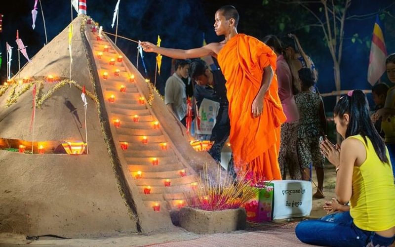 Un moine bouddhiste en robe safran allume des bougies sur un monticule de sable décoré lors des célébrations du Nouvel An cambodgien, tandis qu’une fidèle agenouillée prie à proximité