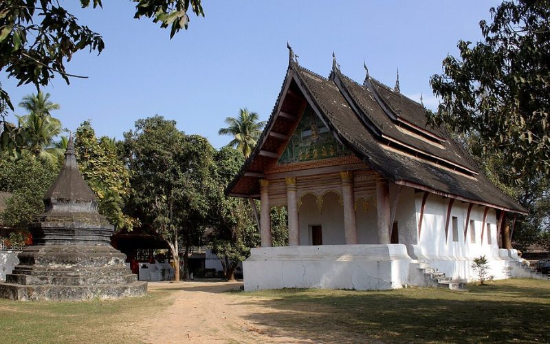 Temple traditionnel laotien à Luang Prabang, entouré de végétation luxuriante, avec un stupa en pierre à proximité, reflétant l'architecture bouddhiste locale