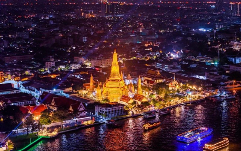Vue aérienne nocturne du Wat Arun illuminé au bord de la rivière Chao Phraya à Bangkok, avec des bateaux éclairés naviguant sur l’eau et la ville brillant en arrière-plan