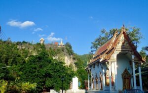 Temple Wat Tha Kha Nun en Thaïlande, entouré de verdure luxuriante, avec une chapelle ornée et des bâtiments perchés sur une colline sous un ciel bleu clair