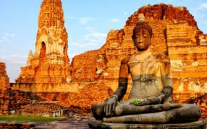 Statue de Bouddha méditant devant les ruines historiques du temple Wat Mahathat à Ayutthaya, illuminées par la lumière dorée du coucher du soleil